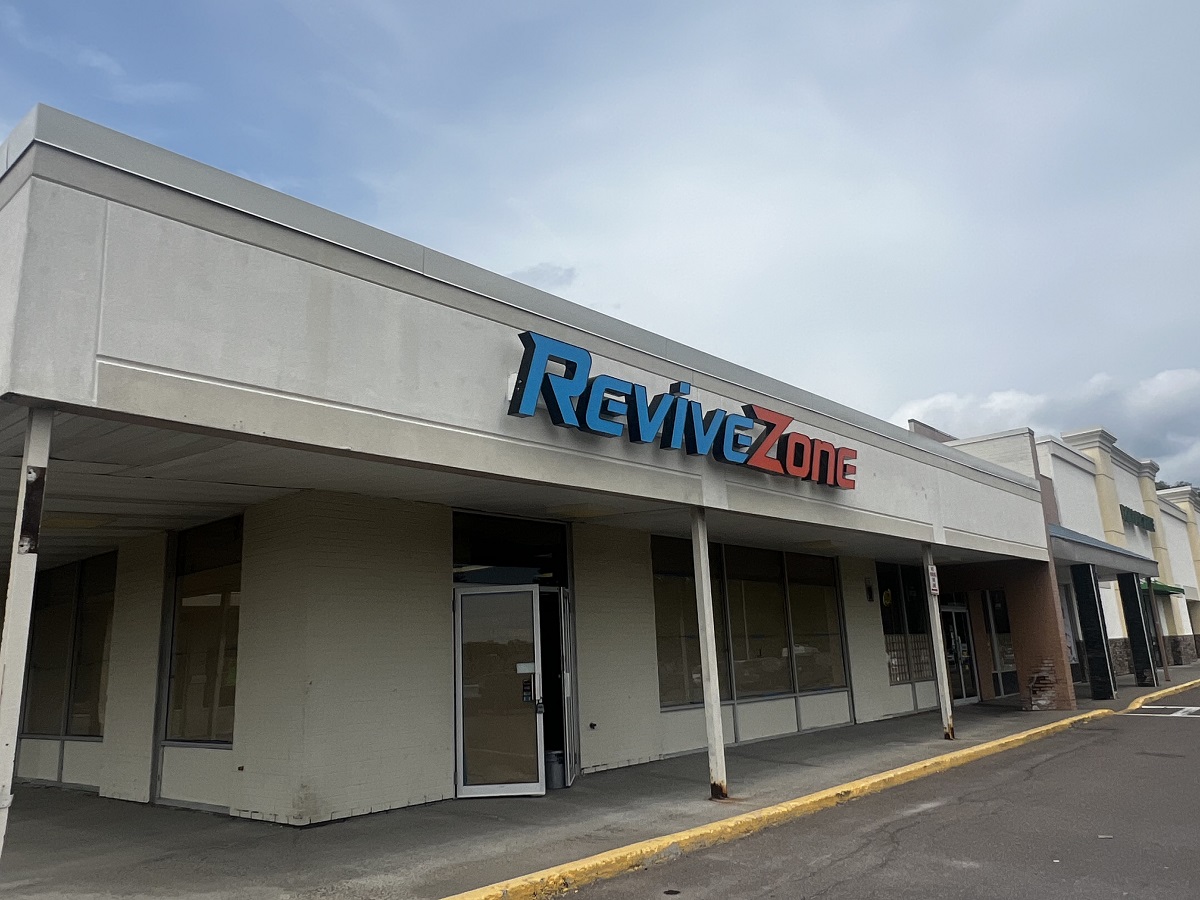 Storefront of a strip mall with a blue and red 'ReviveZone' sign above the entrance, under a pale blue sky