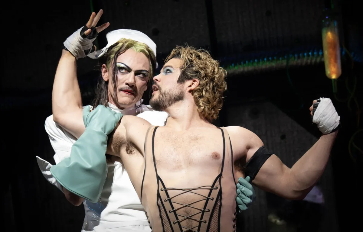 Two performers in theatrical costumes embrace on stage: one in a white nurse outfit with wrapped hands, the other bare-chested in a black lace-up corset, gazing at each other.