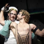 Two performers in theatrical costumes embrace on stage: one in a white nurse outfit with wrapped hands, the other bare-chested in a black lace-up corset, gazing at each other.