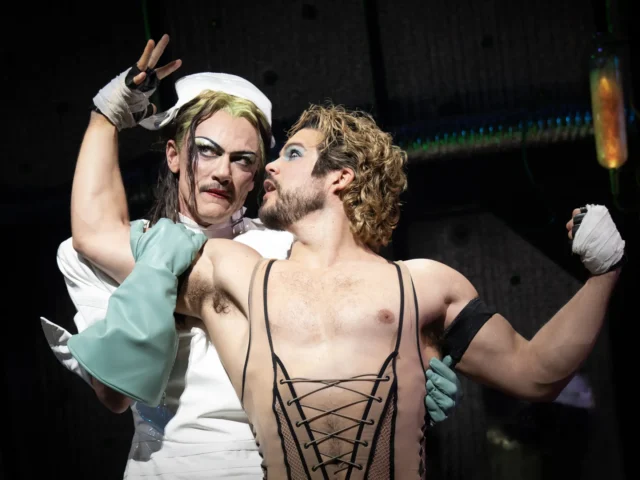 Two performers in theatrical costumes embrace on stage: one in a white nurse outfit with wrapped hands, the other bare-chested in a black lace-up corset, gazing at each other.