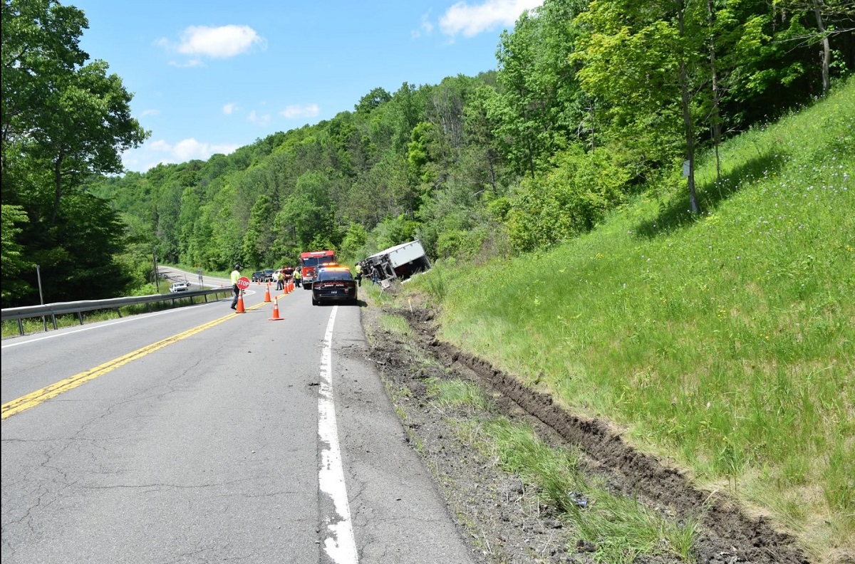 Two-lane rural road scene with a truck off the embankment, emergency vehicles and orange cones blocking the lane during a crash response.