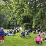 Group of people seated in foldable lawn chairs in a grassy park, watching a small outdoor performance among trees and a wooden fence.