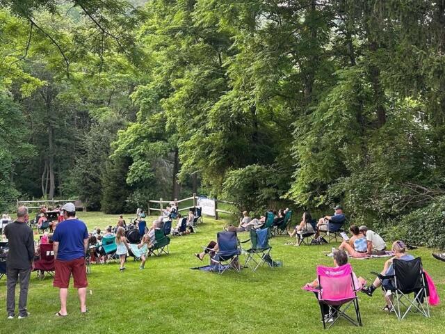 Group of people seated in foldable lawn chairs in a grassy park, watching a small outdoor performance among trees and a wooden fence.