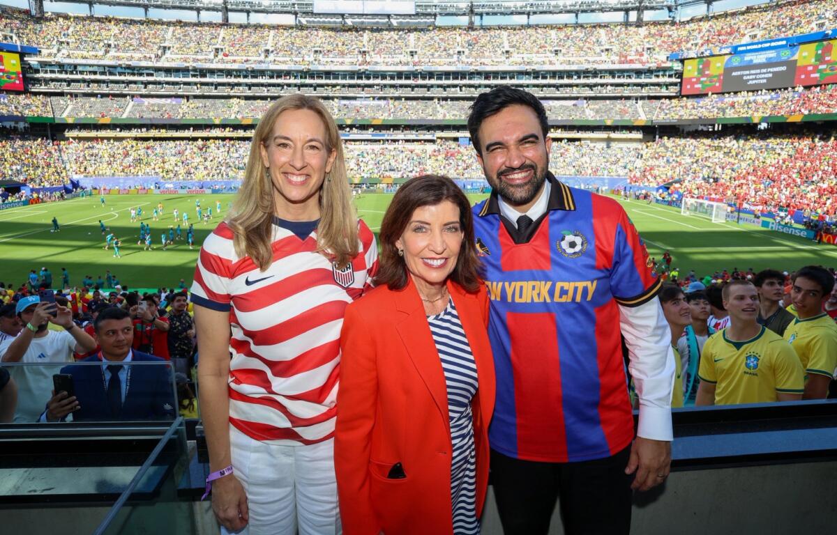 Three smiling fans pose for a photo at a packed soccer stadium, wearing USA-themed jerseys and a red blazer.