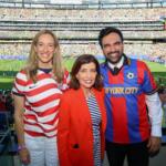Three smiling fans pose for a photo at a packed soccer stadium, wearing USA-themed jerseys and a red blazer.