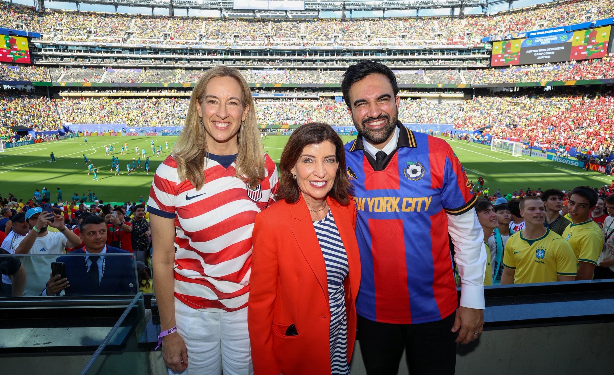 Three smiling fans pose for a photo at a packed soccer stadium, wearing USA-themed jerseys and a red blazer.