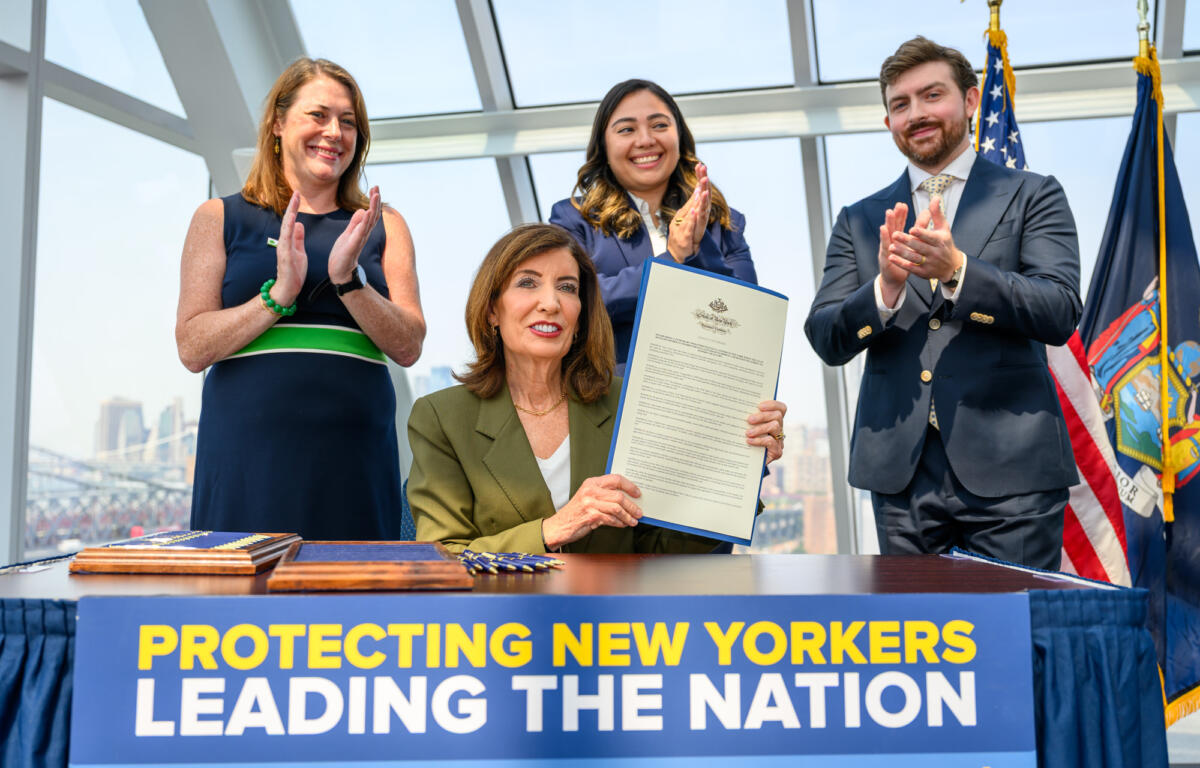 Three officials applaud on stage beside a large banner; a woman in green blazer holds a long document while others clap, with flags in the background and cityscape visible through glass.