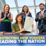 Three officials applaud on stage beside a large banner; a woman in green blazer holds a long document while others clap, with flags in the background and cityscape visible through glass.