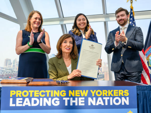 Three officials applaud on stage beside a large banner; a woman in green blazer holds a long document while others clap, with flags in the background and cityscape visible through glass.