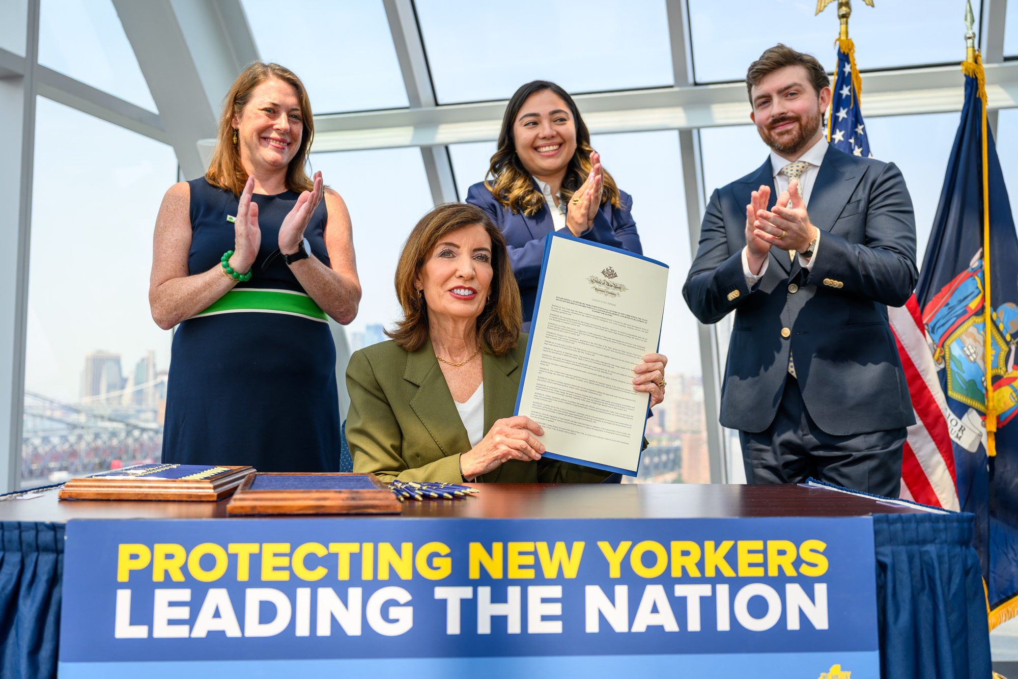 Three officials applaud on stage beside a large banner; a woman in green blazer holds a long document while others clap, with flags in the background and cityscape visible through glass.