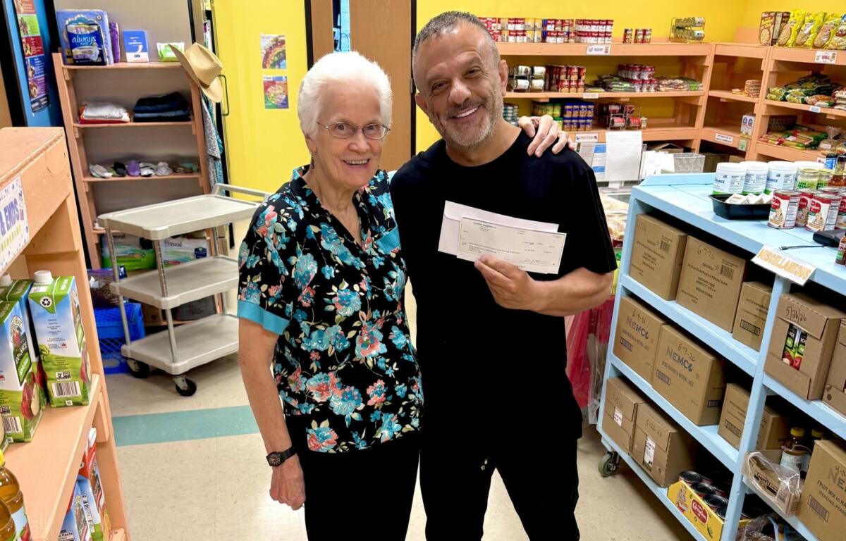 Two smiling adults pose in a pantry holding a large check between them, shelves stocked with groceries in the background.