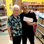 Two smiling adults pose in a pantry holding a large check between them, shelves stocked with groceries in the background.