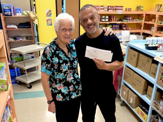 Two smiling adults pose in a pantry holding a large check between them, shelves stocked with groceries in the background.
