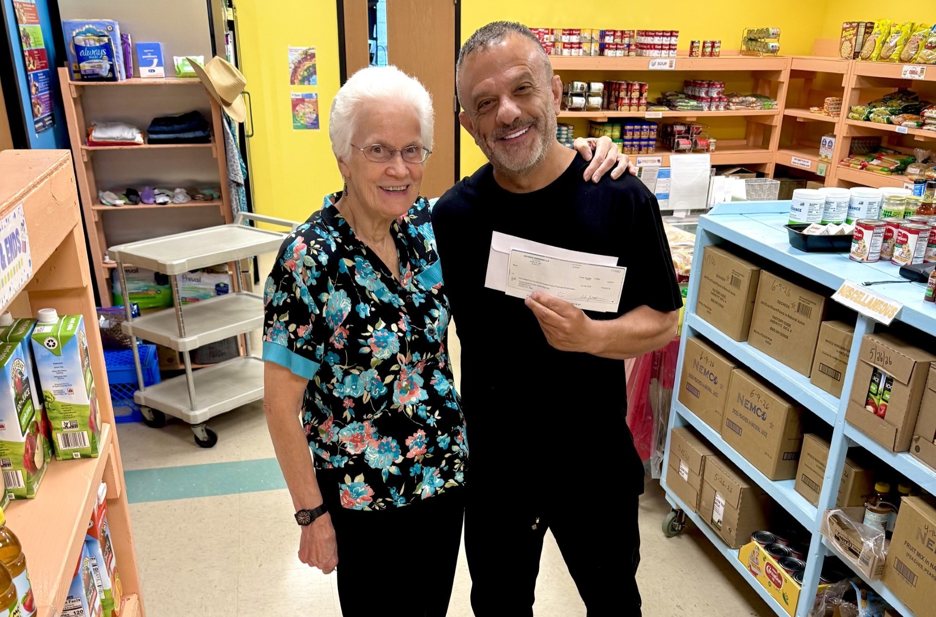 Two smiling adults pose in a pantry holding a large check between them, shelves stocked with groceries in the background.