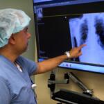 Doctor in blue scrubs and a surgical cap points to a chest X-ray on a wall monitor.