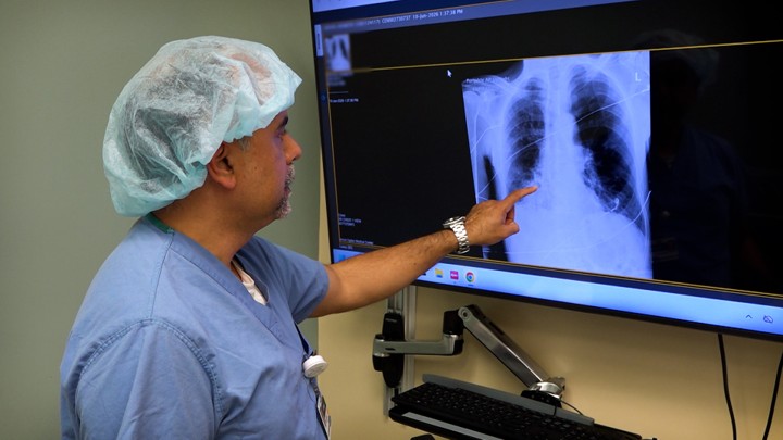 Doctor in blue scrubs and a surgical cap points to a chest X-ray on a wall monitor.