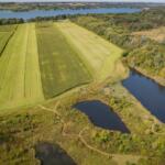 Aerial view of farmland with rectangular green fields, a lake in the distance, and a wetland area with ponds to the right.