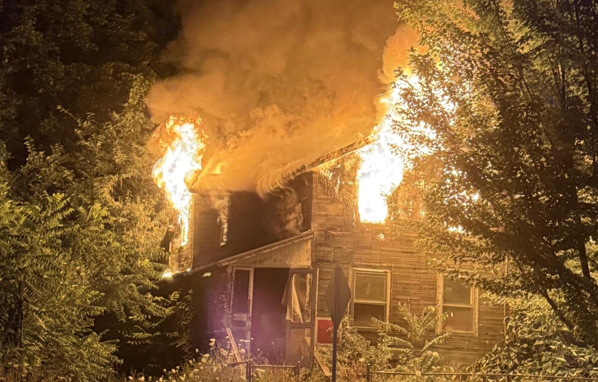 Nighttime house fire: flames engulf the roof and smoke billows upward, surrounded by trees and a front yard fence.