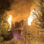 Nighttime house fire: flames engulf the roof and smoke billows upward, surrounded by trees and a front yard fence.