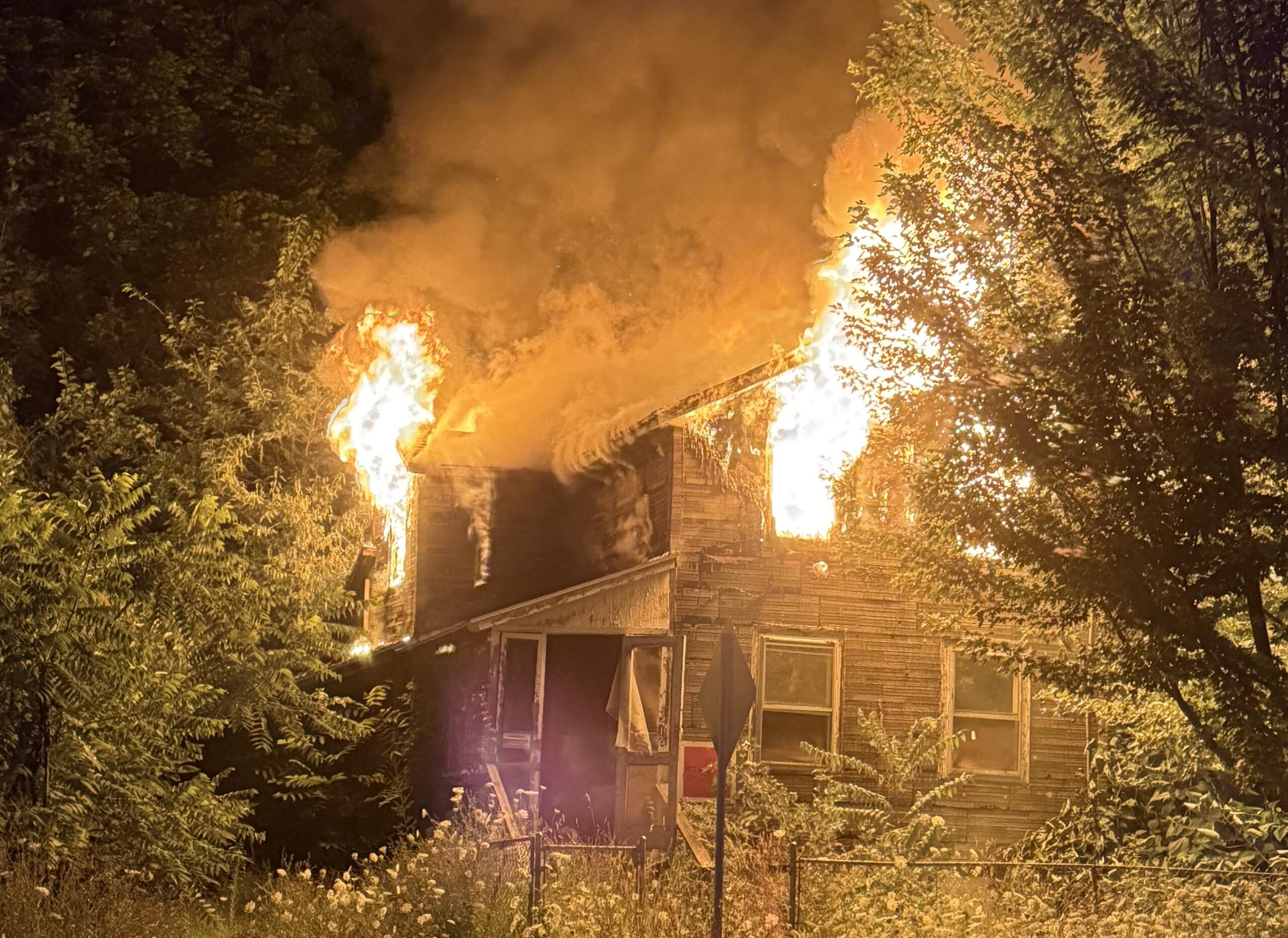 Nighttime house fire: flames engulf the roof and smoke billows upward, surrounded by trees and a front yard fence.