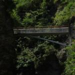 Metal pedestrian bridge spanning a rocky gorge with lush green trees and cliff walls behind it.