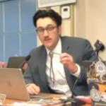 Man in a gray suit speaks into a desk microphone while gesturing, seated at a laptop covered in stickers in a meeting room