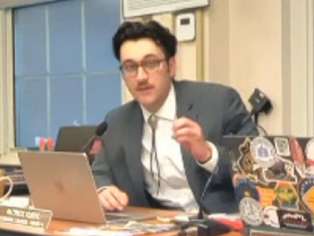 Man in a gray suit speaks into a desk microphone while gesturing, seated at a laptop covered in stickers in a meeting room