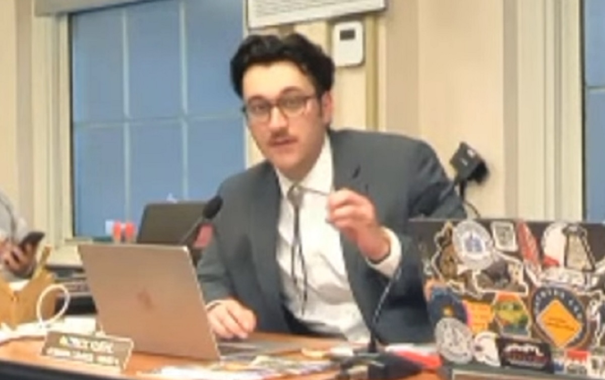 Man in a gray suit speaks into a desk microphone while gesturing, seated at a laptop covered in stickers in a meeting room