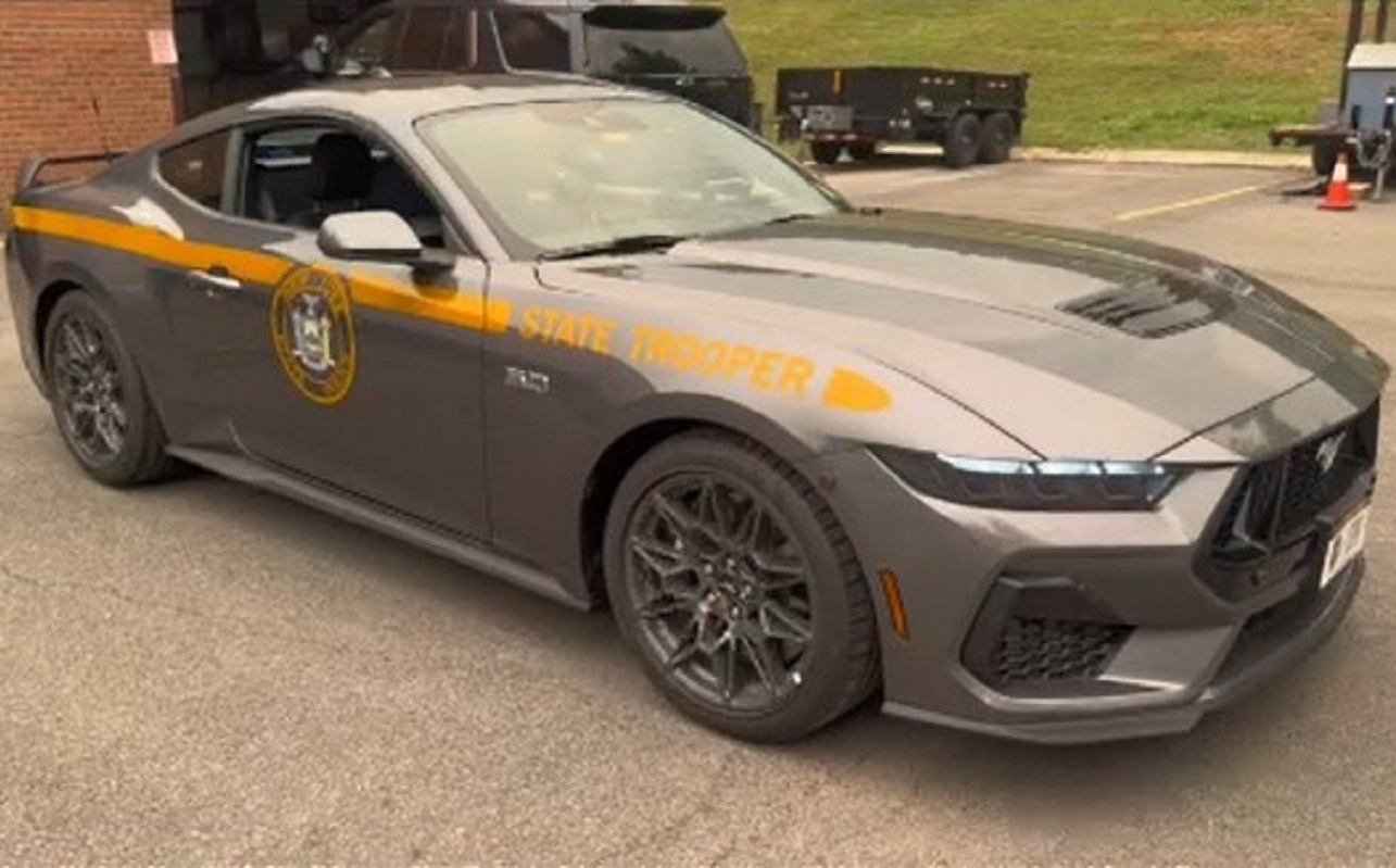 Gray Dodge Charger state trooper patrol car with yellow stripe and emblem parked in a lot on a sunny day.