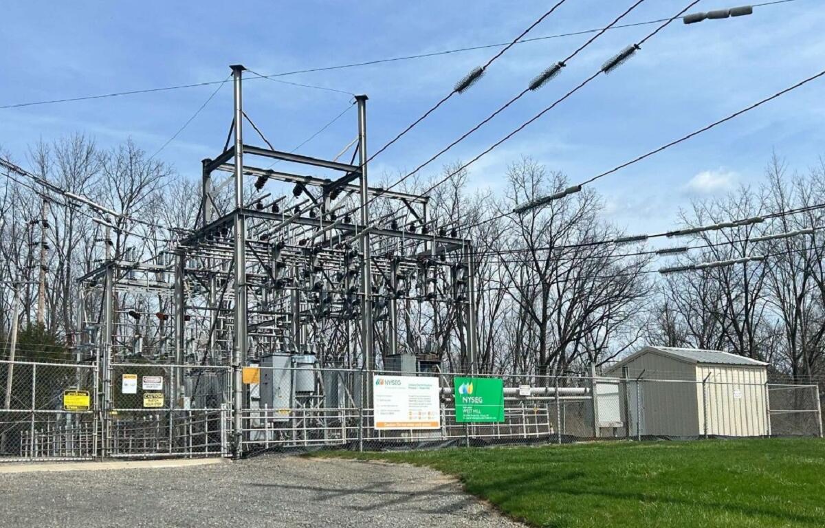 Industrial electric substation behind a chain-link fence with metal framework, transformers, and overhead power lines against a blue sky.