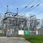 Industrial electric substation behind a chain-link fence with metal framework, transformers, and overhead power lines against a blue sky.