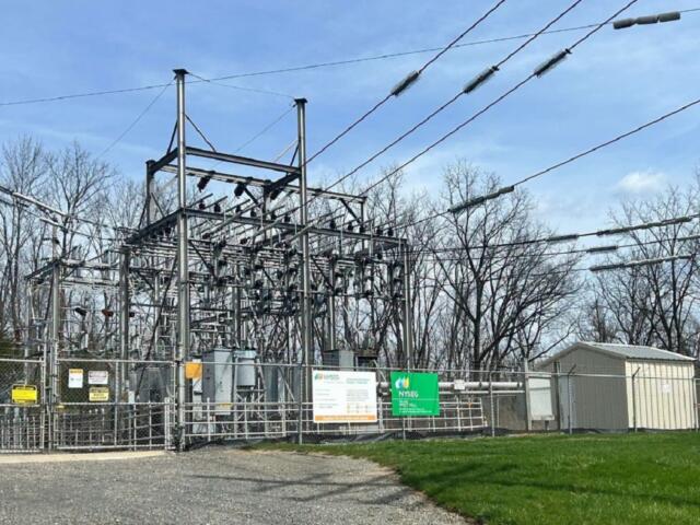 Industrial electric substation behind a chain-link fence with metal framework, transformers, and overhead power lines against a blue sky.