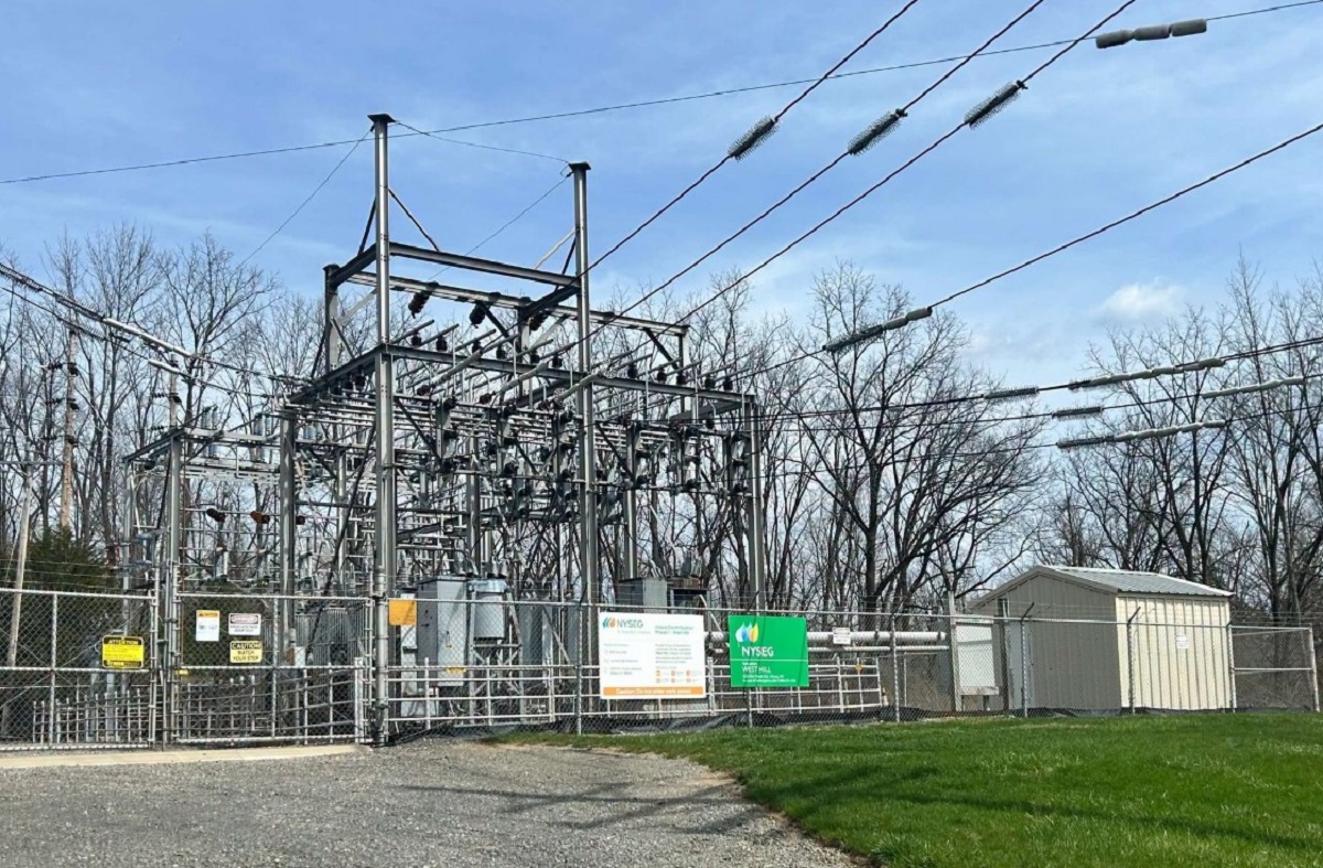 Industrial electric substation behind a chain-link fence with metal framework, transformers, and overhead power lines against a blue sky.