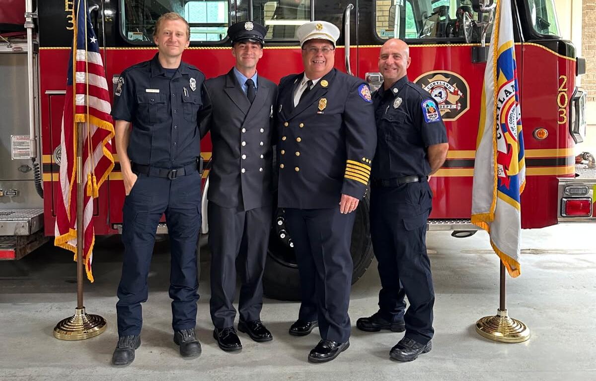 Four uniformed emergency responders pose in front of a red fire truck with flags on either side.