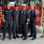 Four uniformed emergency responders pose in front of a red fire truck with flags on either side.