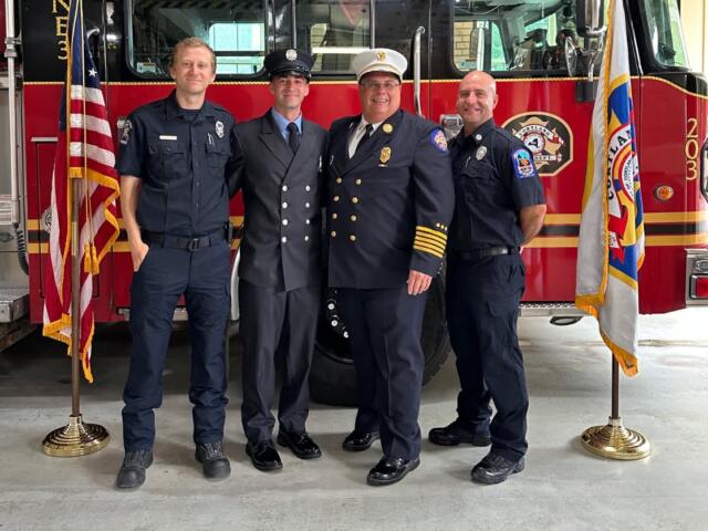 Four uniformed emergency responders pose in front of a red fire truck with flags on either side.