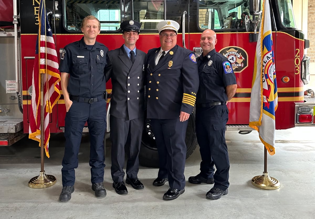 Four uniformed emergency responders pose in front of a red fire truck with flags on either side.