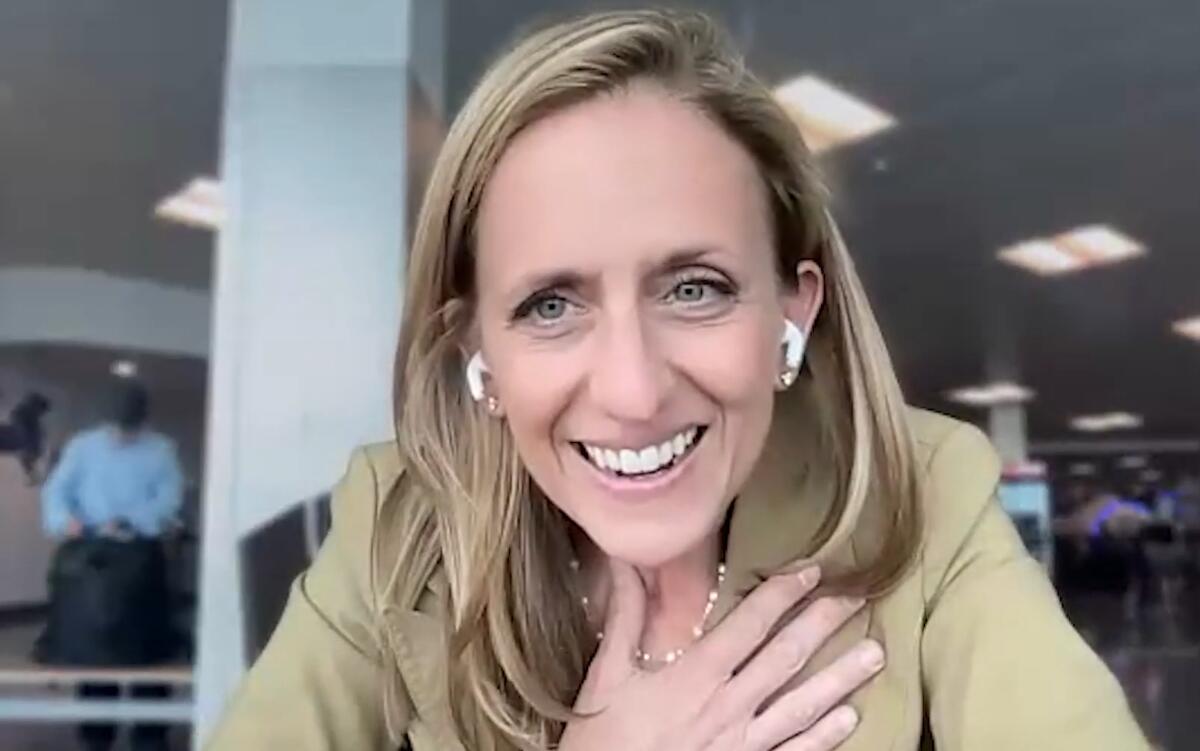 Smiling woman with blond hair wearing wireless earbuds during a video call in an office setting.