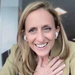 Smiling woman with blond hair wearing wireless earbuds during a video call in an office setting.