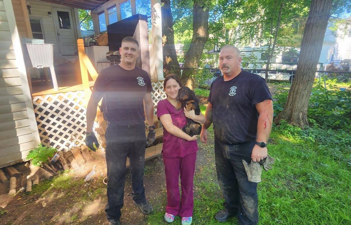 Three adults stand outside a house in a sunny yard; a woman in purple scrubs holds a small dog while two men in dark shirts flank her on either side.