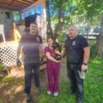 Three adults stand outside a house in a sunny yard; a woman in purple scrubs holds a small dog while two men in dark shirts flank her on either side.