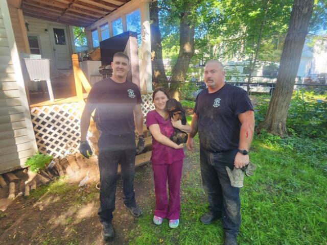 Three adults stand outside a house in a sunny yard; a woman in purple scrubs holds a small dog while two men in dark shirts flank her on either side.