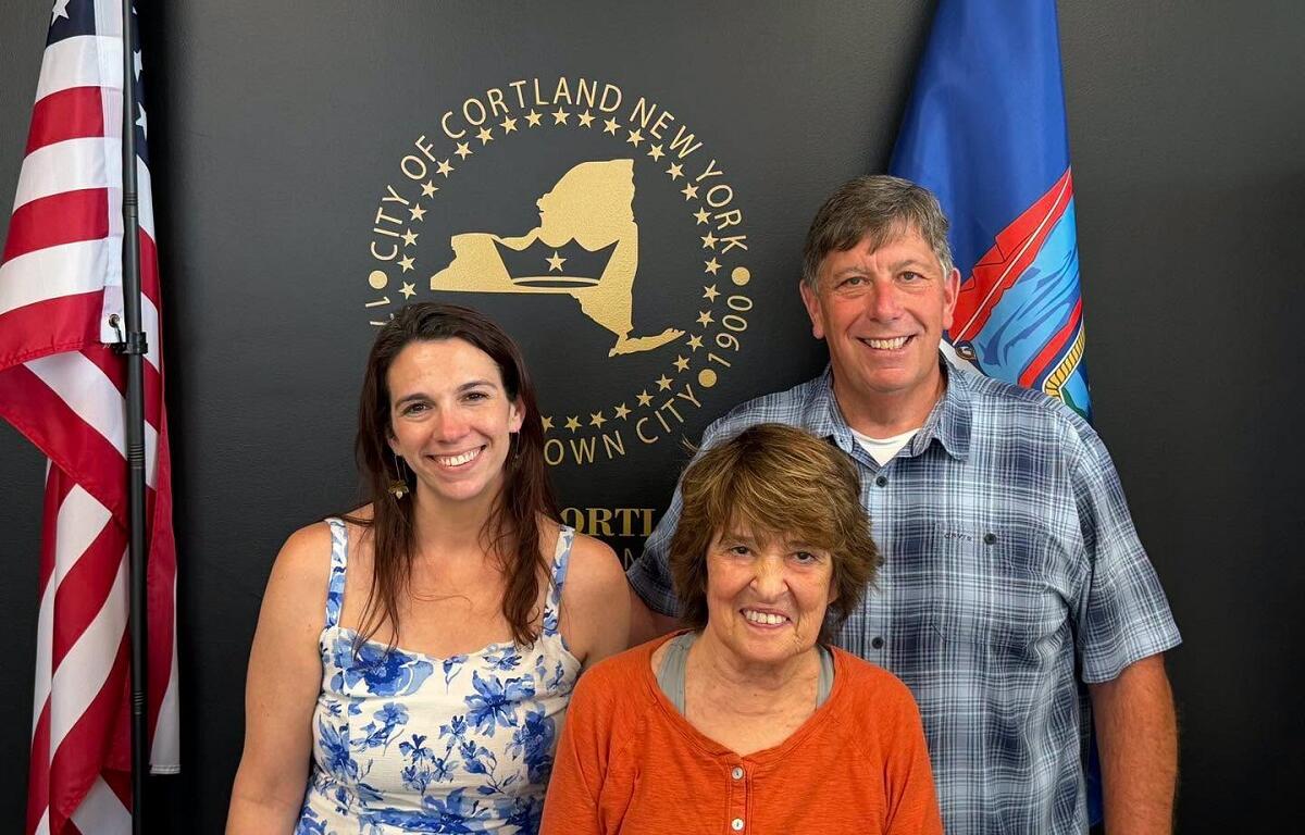 Three adults smiling and posing indoors in front of the Cortland, NY city seal with American and blue flags nearby, backdrop dark wall.