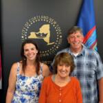 Three adults smiling and posing indoors in front of the Cortland, NY city seal with American and blue flags nearby, backdrop dark wall.