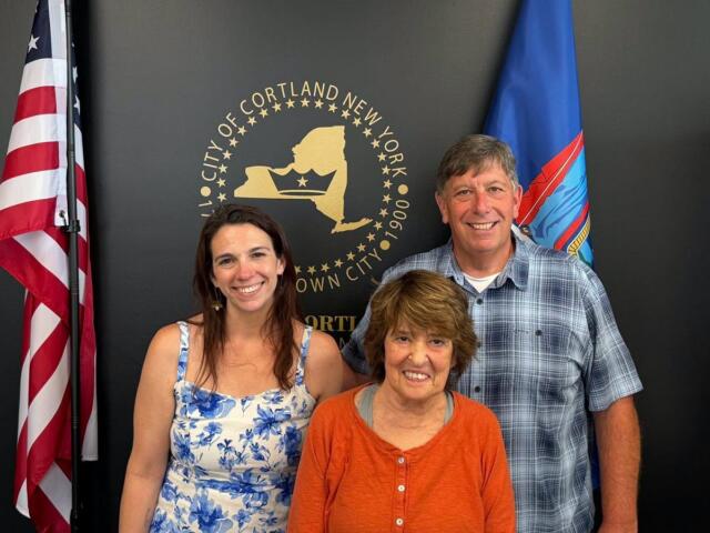 Three adults smiling and posing indoors in front of the Cortland, NY city seal with American and blue flags nearby, backdrop dark wall.