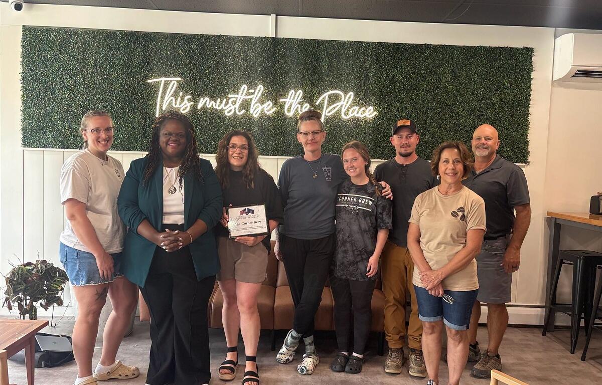 Group portrait of nine adults indoors in front of a green hedge wall with a neon sign that reads 'This must be the Place'; one person holds a certificate and everyone smiles.