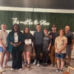 Group portrait of nine adults indoors in front of a green hedge wall with a neon sign that reads 'This must be the Place'; one person holds a certificate and everyone smiles.