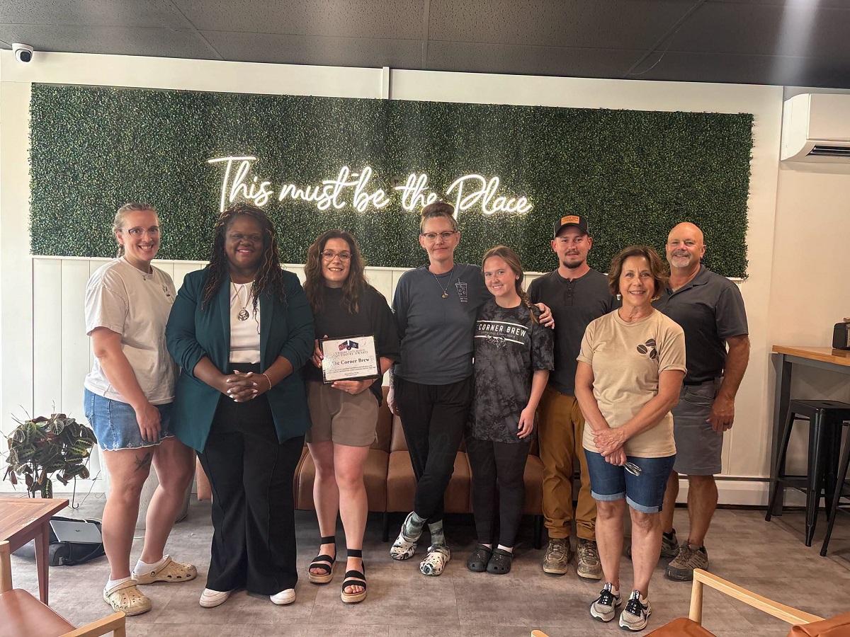 Group portrait of nine adults indoors in front of a green hedge wall with a neon sign that reads 'This must be the Place'; one person holds a certificate and everyone smiles.