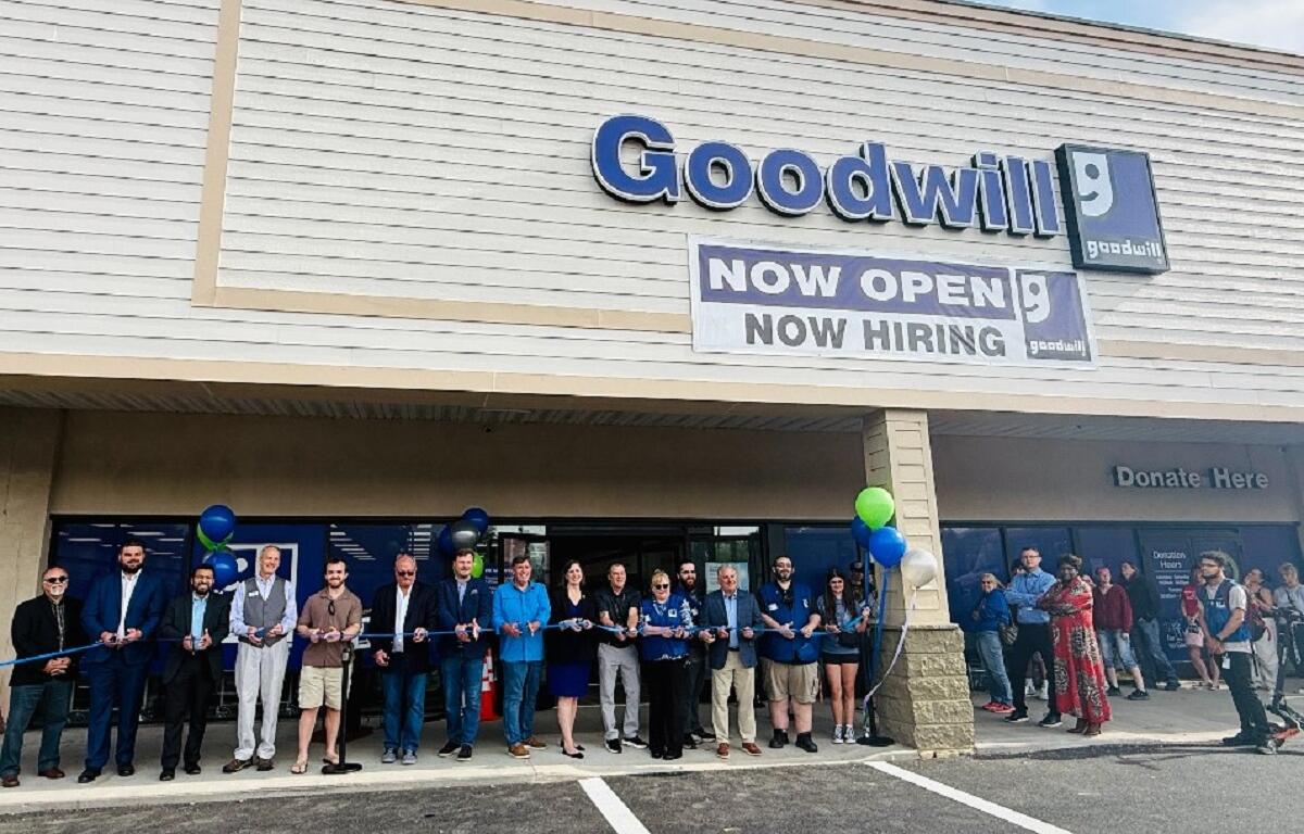 Group of people cutting a blue ribbon outside a Goodwill store during a grand opening event with balloons nearby.