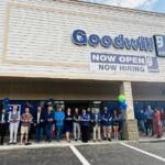 Group of people cutting a blue ribbon outside a Goodwill store during a grand opening event with balloons nearby.